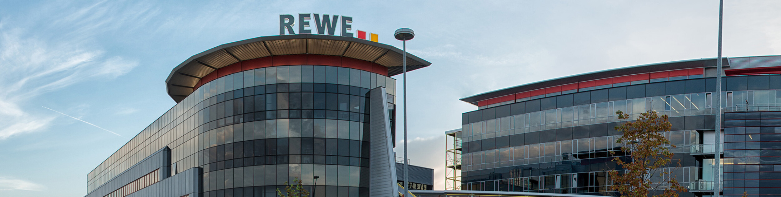Moderne Bürogebäude mit dem Logo "REWE" auf dem Dach. Die Fassade besteht aus Glas und Metall.