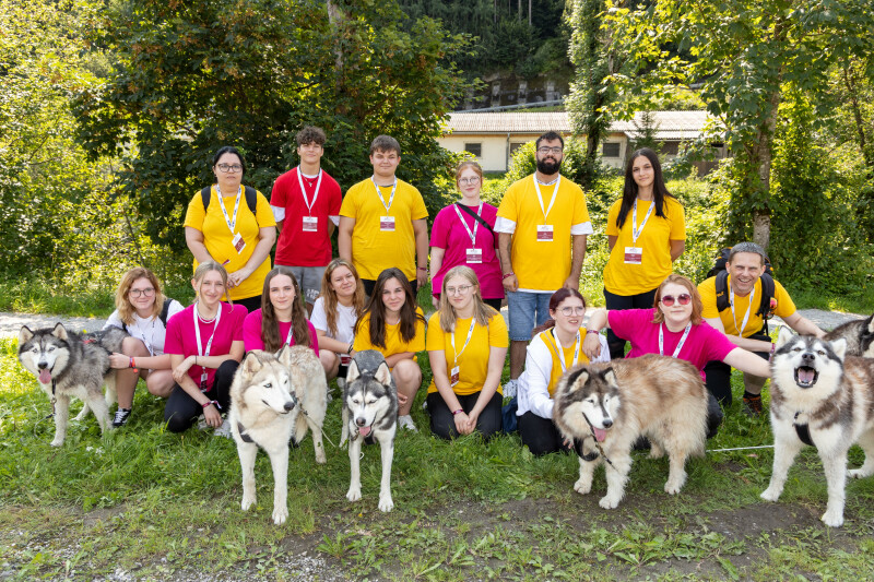 Eine Gruppe von Menschen steht und kniet im Freien neben mehreren Huskys. Die Personen tragen gelbe und pinke T-Shirts und haben Namensschilder um den Hals. Im Hintergrund sind Bäume und ein Gebäude zu sehen.