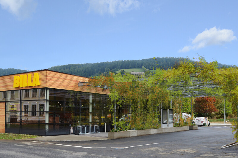 Ein Supermarkt mit der Aufschrift "BILLA" an der Fassade, umgeben von Vegetation und einer überdachten Struktur mit Pflanzen. Im Hintergrund sind Hügel und ein blauer Himmel zu sehen.