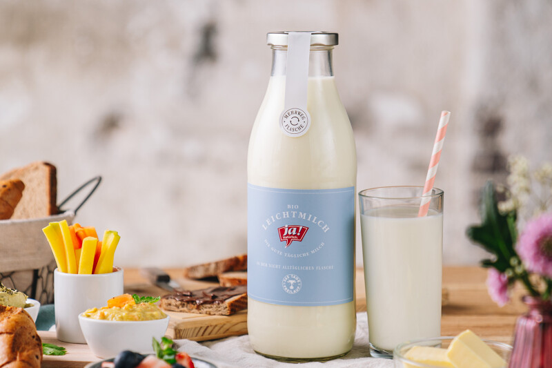 Zwei Flaschen Bio-Frischmilch auf einem Tisch, begleitet von einem Glas Milch, Brot, Aufstrichen und einem Blumentopf. Im Hintergrund unscharfe Brotkorb und Snacks.