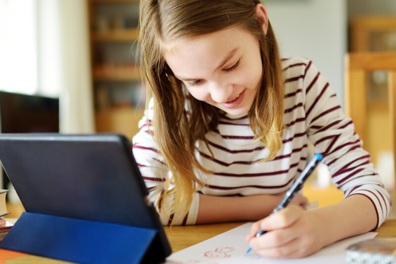 Ein Mädchen mit gestreiftem Pullover schreibt etwas auf ein Blatt Papier und blickt dabei auf ein Tablet. Sie sitzt an einem Tisch in einem Raum mit Bücherregal im Hintergrund.