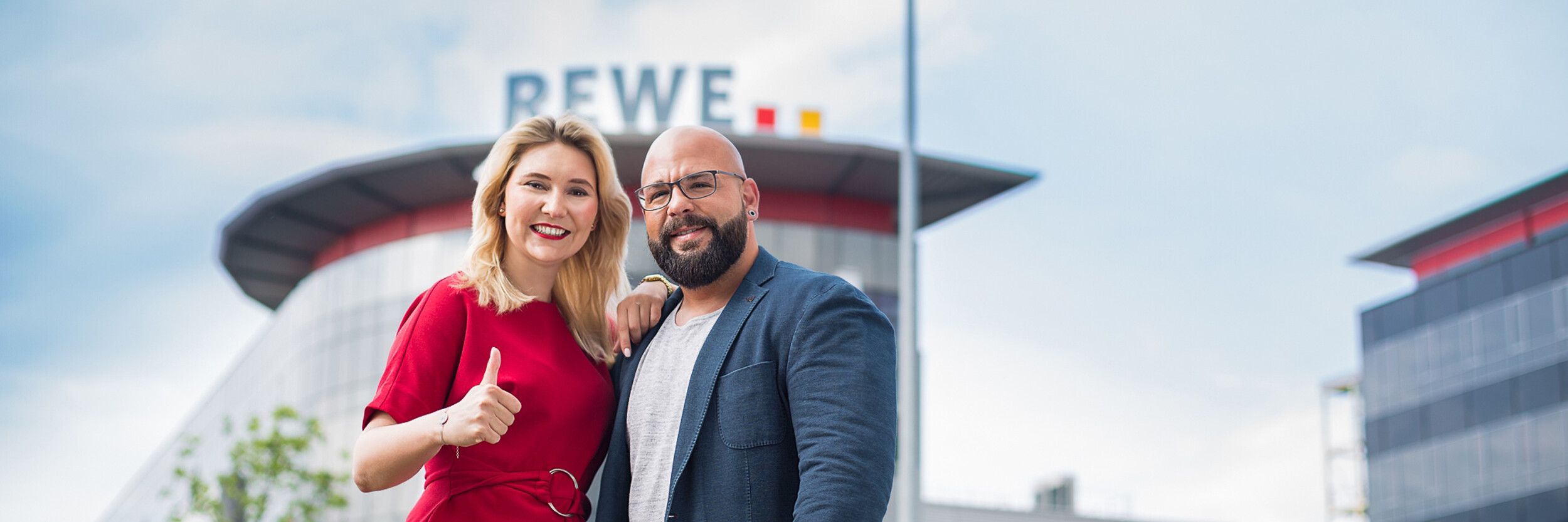 Zwei Personen stehen lächelnd vor einem modernen Gebäude mit dem Schriftzug "REWE" auf dem Dach. Die Frau trägt ein rotes Kleid, der Mann einen dunklen Anzug und ein weißes Hemd. Sie zeigt den Daumen nach oben.