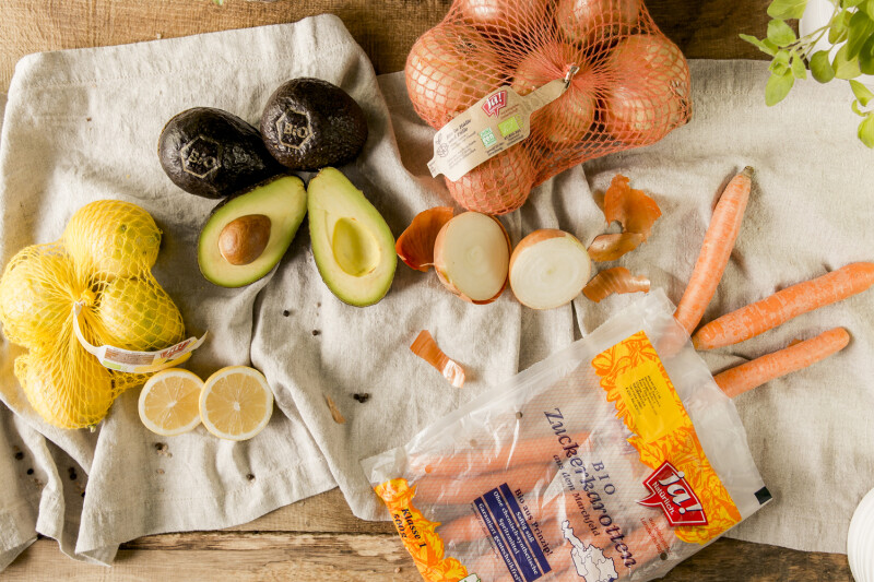 Obst und Gemüse auf einem Leinentuch, darunter Avocados, Zitronen, Möhren, Zwiebeln und ein Beutel Zuckermöhren. Eine Pflanze ist am Bildrand sichtbar.