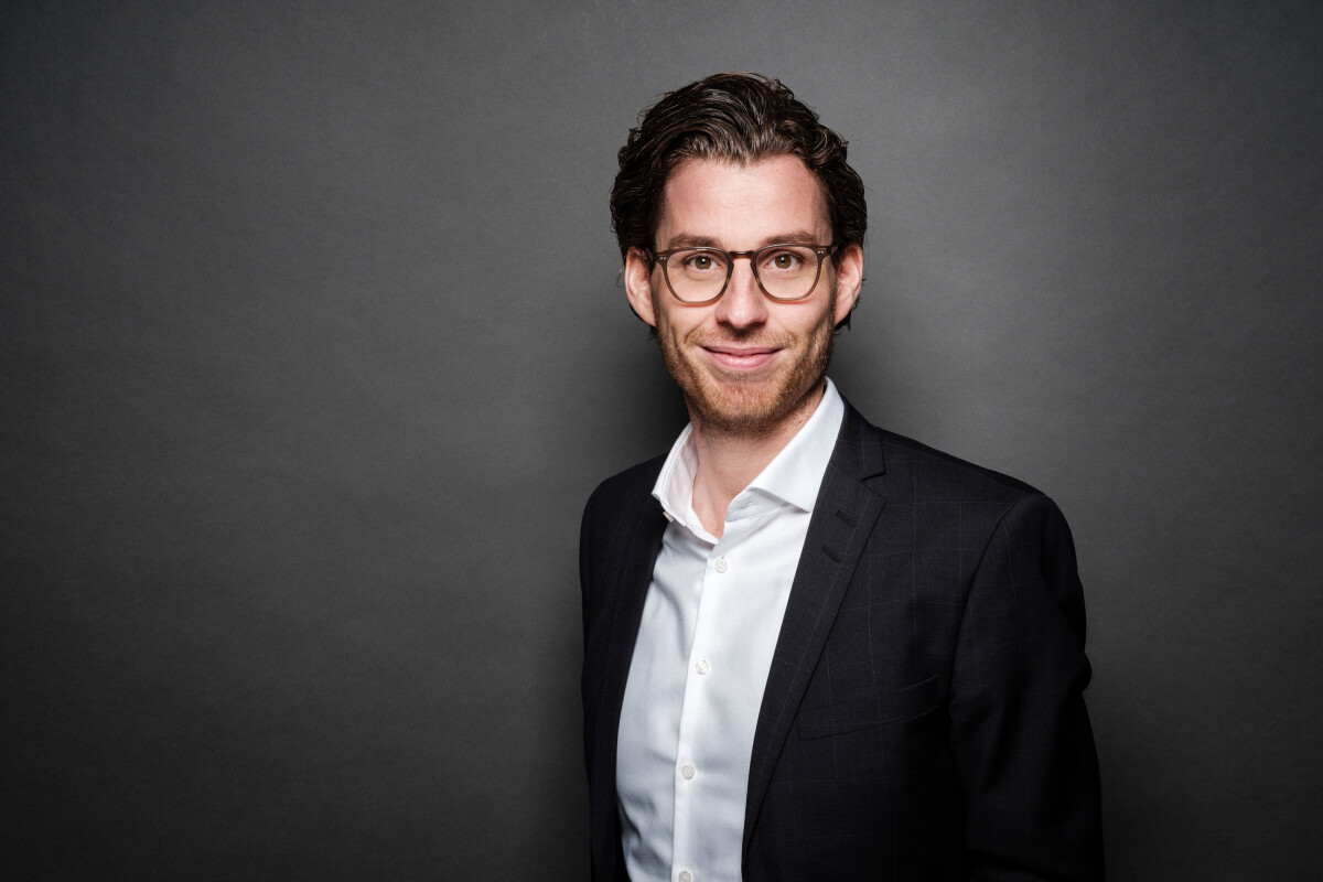 Ein lächelnder Mann mit Brille, dunklem lockigem Haar und einem dunklen Anzug vor einem grauen Hintergrund.