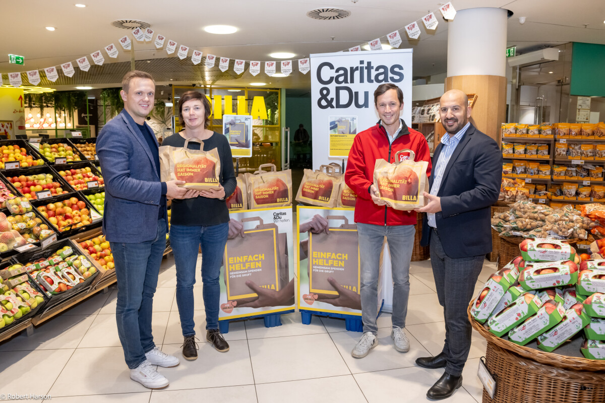 Vier Personen stehen in einem Supermarkt vor einem Stand mit Caritas- und Billa-Taschen. Zwei Männer und eine Frau halten Einkaufstaschen. Im Hintergrund sind Obstkörbe und Werbeplakate zu sehen.