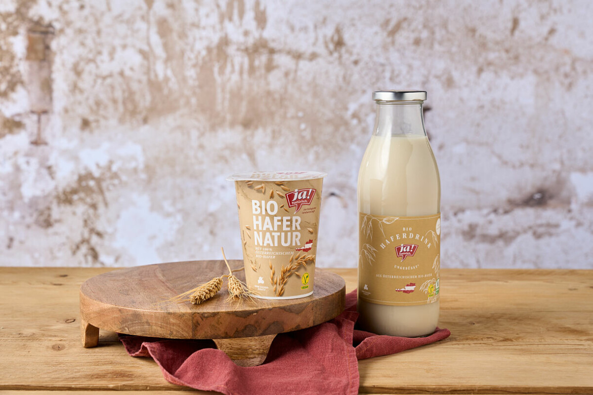 Ein Becher Bio-Haferjoghurt und eine Glasflasche Hafermilch stehen auf einem hölzernen Tisch vor einer rustikalen Wand. Daneben liegt ein Ährenzweig und ein rotes Tuch.