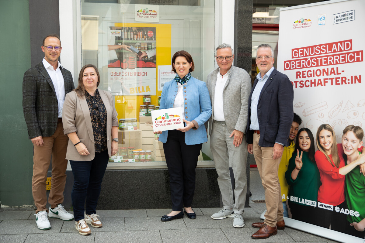 Mehrere Personen stehen vor einem Geschäft neben einem Banner mit der Aufschrift „Genussland Oberösterreich Regionalbotschafter:in“. Sie lächeln und halten Schilder in der Hand.