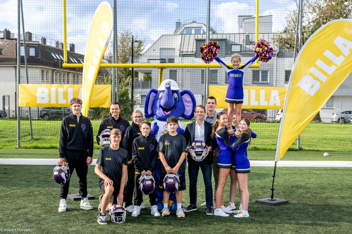 Eine Gruppe von jungen Sportlern und Cheerleadern posiert auf einem Fußballfeld. Im Hintergrund steht ein maskottchenartiger blauer Elefant. Auf den gelben Fahnen steht "BILLA". Im Hintergrund sind Gebäude und ein torähnliches Konstrukt zu sehen.