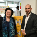 Eine Frau und ein Mann stehen lächelnd in einem Raum. Im Hintergrund sind dekorierte Fenster und ein gelber Banner mit einem Logo und der Aufschrift "Hilfswerk".