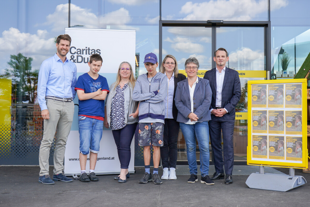 Eine Gruppe von sieben Personen steht vor einem Gebäude mit Glasfront. Hinter ihnen ist ein Schild von "Caritas & Du" zu sehen. Alle tragen legere Kleidung, einige haben die Arme verschränkt. Rechts steht eine Infotafel mit Plakaten. Der Himmel ist bewölkt.