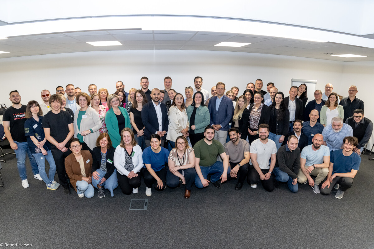 Große Gruppe von Menschen, die in einem Konferenzraum posieren. Die Personen stehen und sitzen in mehreren Reihen und lächeln in die Kamera. Im Hintergrund ist eine weiße Wand mit einem Flipchart sichtbar.