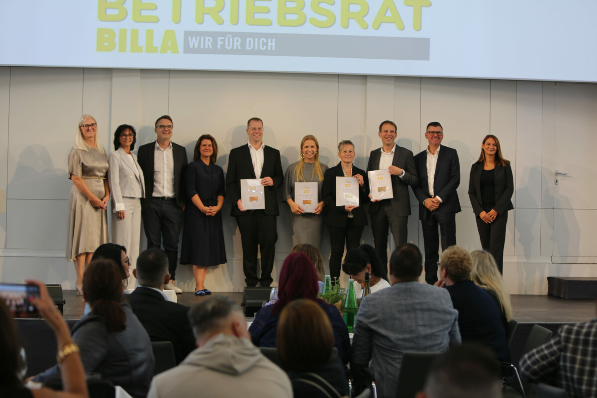 Eine Gruppe von Männern und Frauen in formeller Kleidung steht auf einer Bühne und hält Dokumente. Über ihnen ist ein Schild mit der Aufschrift "Betriebsrat Billa Wir Für Dich" zu sehen. Vor der Bühne sitzen mehrere Zuschauer.