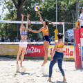 Zwei Frauen spielen Beachvolleyball auf einem Sandplatz. Eine Spielerin springt, um den Ball über das Netz zu schlagen, während die andere versucht, ihn zu blocken. Der Schiedsrichter beobachtet das Spiel von seinem Stand aus. Im Hintergrund sind Bäume und Werbebanner zu sehen.