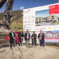 Fünf Männer in Anzügen mit Schaufeln stehen vor einer Baustelle mit einem großen Bauschild und einem Bagger im Hintergrund. Ein Banner mit einem Bauprojekt ist ebenfalls zu sehen. Im Hintergrund Berglandschaft.