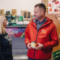 Ein Mann und eine Frau unterhalten sich in einem Lebensmittelgeschäft. Der Mann trägt eine rote Jacke mit einem Logo, die Frau trägt einen schwarzen Pullover mit einem Schriftzug. Sie stehen zwischen Regalen mit verpackten Lebensmitteln und frischem Obst.
