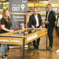 Vier Personen stehen um einen Tischkicker in einem Lebensmittelladen. Links eine Frau mit schwarzem T-Shirt, daneben drei Männer in Anzügen. Im Hintergrund sind Regale mit Produkten zu sehen. Ein Banner mit der Aufschrift "Tischlein Check Dich" steht daneben.