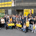 Eine Gruppe von Menschen versammelt sich vor einem Gebäude mit der Aufschrift "Ausbildungszentrum Billa". Einige halten gelbe Schilder mit Text. Die Stimmung wirkt fröhlich und gemeinsam. Im Hintergrund sind große Fenster und Pflanzen zu sehen.