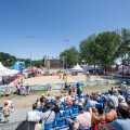 Eine Beachvolleyball-Veranstaltung mit einem Match auf einem sandigen Spielfeld. Zuschauer sitzen auf blauen Tribünenstühlen. Im Hintergrund sind Zelte, ein großer Bildschirm und Bäume zu sehen. Der Himmel ist klar und sonnig.