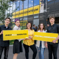 Fünf junge Menschen stehen lächelnd vor einem Gebäude mit Glasfront und halten Schilder mit der Aufschrift "#NEXTBILLAGENERATION". Im Hintergrund sind weitere Personen und ein gelbes Banner mit der Aufschrift "AUSBILDUNGSZENTRUM" zu sehen.