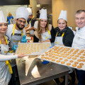 Eine Gruppe von Menschen in Kochuniformen und weißen Hüten präsentiert frische Plätzchen auf Backblechen in einer Küche. Alle lächeln in die Kamera.
