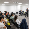 Eine Gruppe von Personen sitzt in einem Konferenzraum und hört einem Mann im Anzug zu, der spricht. Neben ihm steht eine Frau, und im Hintergrund sitzen zwei weitere Männer an einem Tisch.