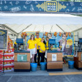 Vier Personen in gelben T-Shirts stehen vor einem Billa Food Stand auf einem Festival. Hinter ihnen sind Verkaufsstände mit Snacks und Getränken. Der Stand ist mit einem Billa-Logo und dekorativen Musiknoten versehen.