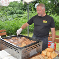 Ein Mann grillt Fleisch auf einem großen Grill im Freien. Im Vordergrund sind Brötchen zusehen. Im Hintergrund ist grüne Vegetation. Der Mann trägt ein schwarzes T-Shirt und benutzt Grillzangen.