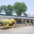 Weißes Festzelt mit spitzen Dächern, umgeben von Menschen. Im Vordergrund stehen gelbe Liegestühle und Sonnenschirme. Einige Besucher gehen auf einem Kiesweg, während andere auf der Holzterrasse des Zelts stehen. Im Hintergrund sind Bäume und blauer Himmel.