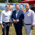 Drei Männer stehen auf einer Wiese und lächeln in die Kamera. Im Hintergrund sind ein gelbes Werbeplakat und rote Zelte zu sehen.