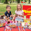Zwei lächelnde Kinder sitzen auf einer Picknickdecke im Park, umgeben von verschiedenen Lebensmitteln und Getränken. Im Hintergrund ist ein großes Schild mit PENNY MARKT-Logo zu sehen.