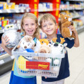 Zwei lächelnde Kinder stehen in einem Supermarktgang und halten einen Einkaufskorb voller Plüschtiere, darunter ein Eisbär, ein Rentier und eine Eule. Regale mit weiteren Produkten befinden sich im Hintergrund.