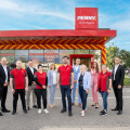 Eine Gruppe von Menschen steht vor einem PENNY-Markt mit einem Eingang, der mit roten und gelben Luftballons dekoriert ist. Die Personen tragen größtenteils rote Kleidung mit dem PENNY-Logo, während einige in formeller Kleidung gekleidet sind. Der Himmel ist blau mit einigen Wolken im Hintergrund.