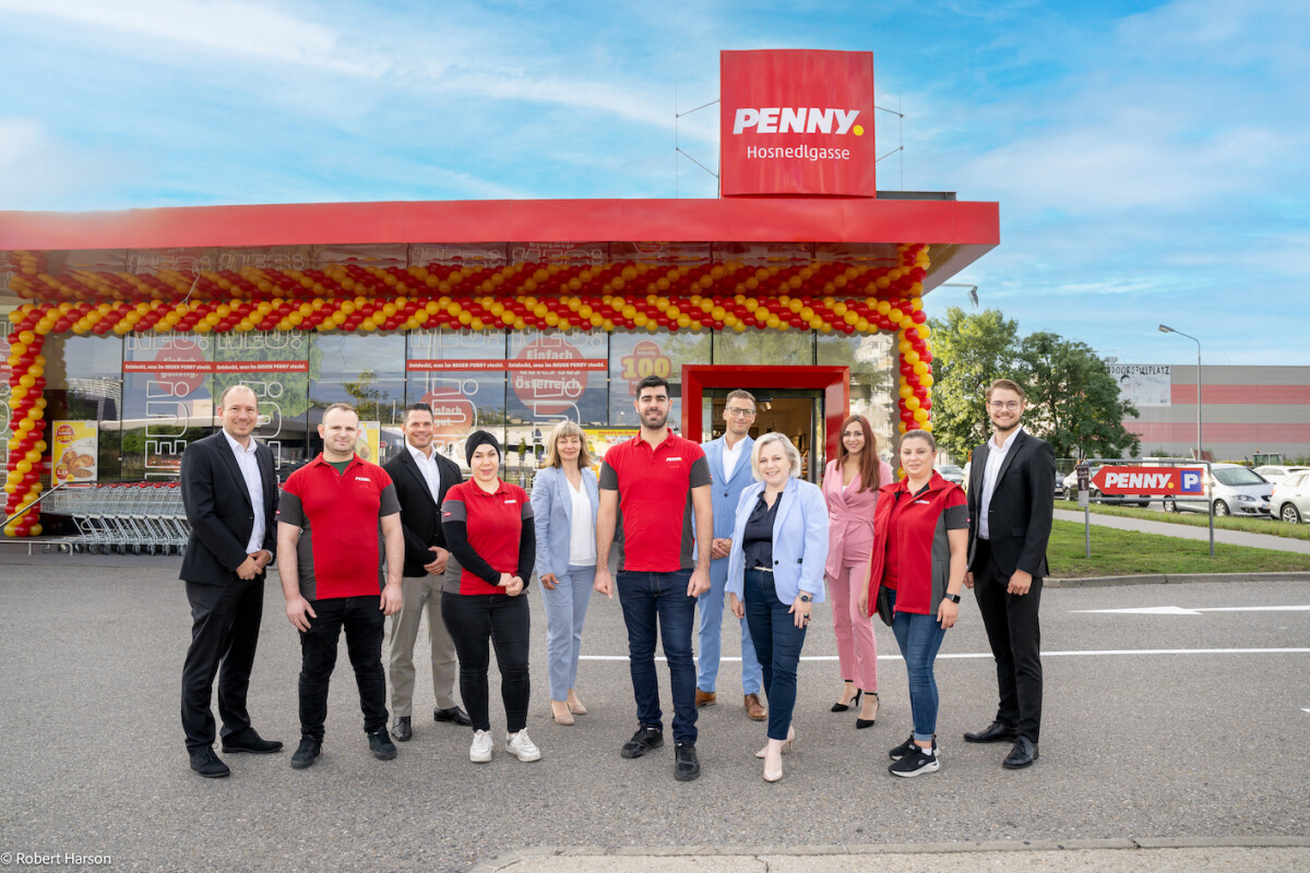 Eine Gruppe von Menschen steht vor einem PENNY-Markt mit einem Eingang, der mit roten und gelben Luftballons dekoriert ist. Die Personen tragen größtenteils rote Kleidung mit dem PENNY-Logo, während einige in formeller Kleidung gekleidet sind. Der Himmel ist blau mit einigen Wolken im Hintergrund.