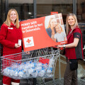 Zwei Frauen in roter Kleidung stehen vor einem Einkaufswagen voller Wasserflaschen. Sie halten ein Schild mit der Aufschrift "PENNY hilft Familien in Not in Österreich" und dem Logo des Roten Kreuzes. Im Hintergrund sind eine Glasfassade und Eingangstüren zu sehen.