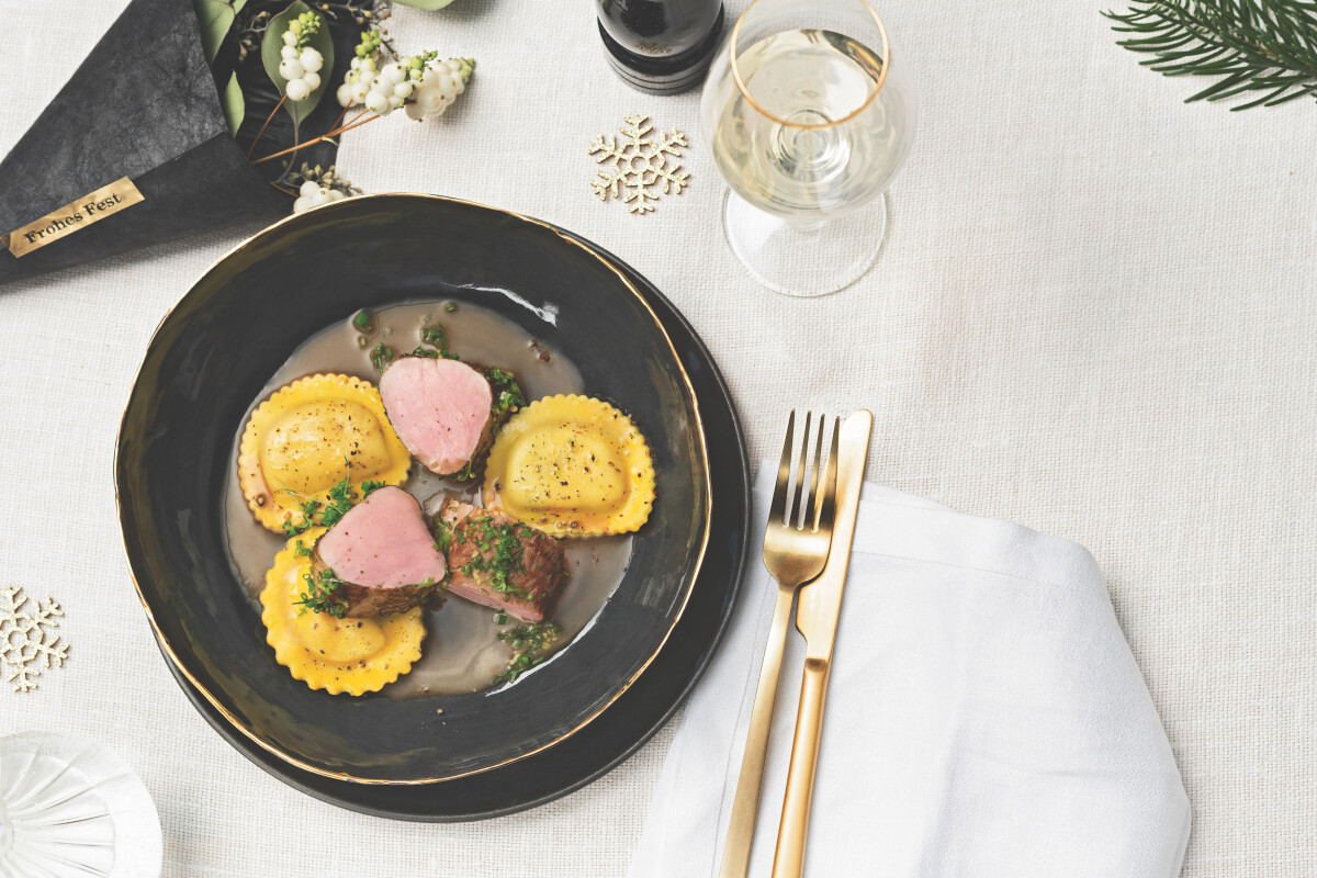 Ein eleganter Teller mit gelben Ravioli, zwei Stück Schweinefilet und einer dunklen Sauce, daneben ein Glas Weißwein und goldfarbenes Besteck auf einem weißen Tisch mit schlichter Dekoration.