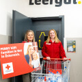 Zwei Frauen stehen vor einer Tür mit der Aufschrift "Leergut". Eine Frau hält ein Schild mit dem Text "PENNY hilft Familien in Not in Österreich". Die andere Frau steht neben einem Einkaufswagen voller leerer Plastikflaschen. Beide lächeln.