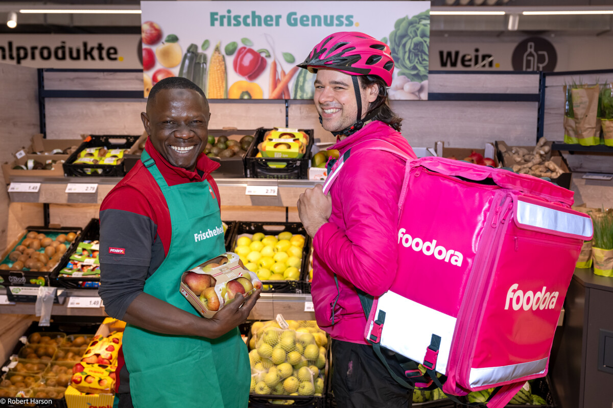 Ein lächelnder Kassierer in grüner Schürze hält einen Sack mit Äpfeln. Daneben steht ein Foodora-Lieferfahrer in pinker Jacke und Helm mit einem großen pinkfarbenen Liefer-Rucksack. Im Hintergrund sind frisches Obst und Gemüse sowie Schilder mit der Aufschrift "Frischer Genuss".