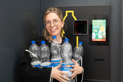 Eine Person steht vor einem Pfandautomaten und hält mehrere leere Plastikflaschen in den Armen. Sie trägt eine Brille und lächelt. Im Hintergrund ist ein Bildschirm mit Anweisungen zur Pfandrückgabe zu sehen.