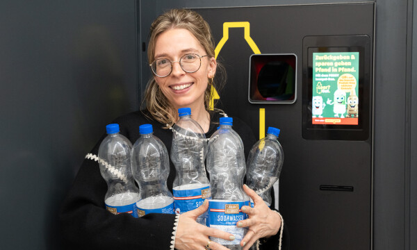 Eine Person steht vor einem Pfandautomaten und hält mehrere leere Plastikflaschen in den Armen. Sie trägt eine Brille und lächelt. Im Hintergrund ist ein Bildschirm mit Anweisungen zur Pfandrückgabe zu sehen.