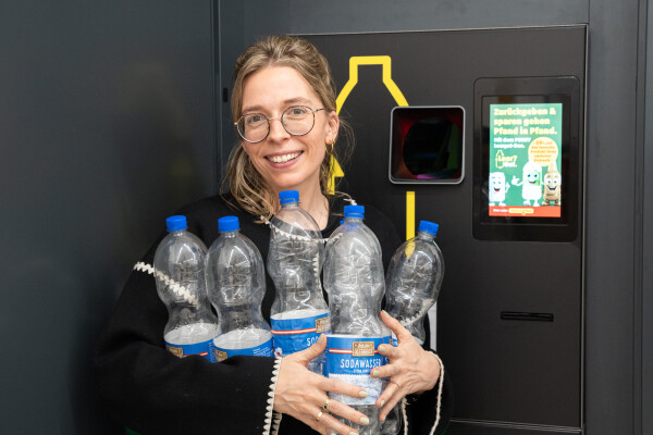 Eine Person steht vor einem Pfandautomaten und hält mehrere leere Plastikflaschen in den Armen. Sie trägt eine Brille und lächelt. Im Hintergrund ist ein Bildschirm mit Anweisungen zur Pfandrückgabe zu sehen.
