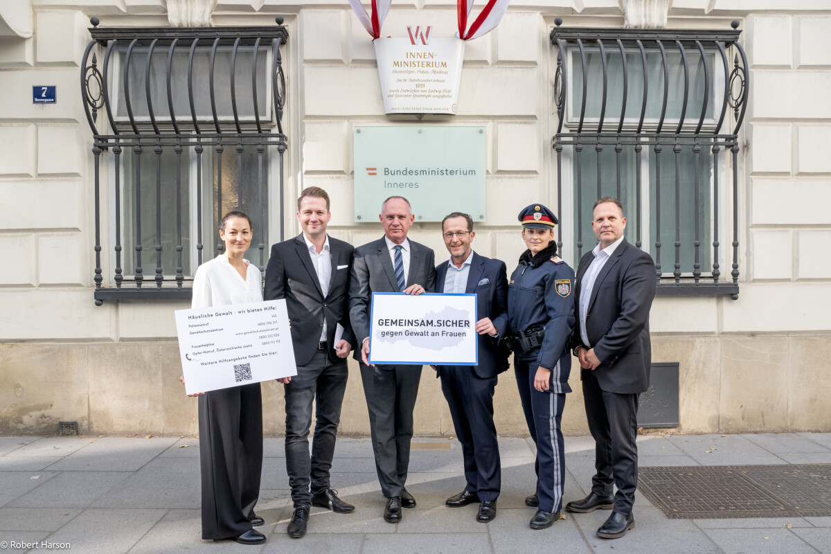 Sechs Personen stehen vor einem Gebäude mit dem Schild "Bundesministerium Inneres". Eine Person hält ein Schild mit der Aufschrift "GEMEINSAMSICHER" und eine andere einen QR-Code. Eine der Personen trägt eine Polizeiuniform. Alle lächeln in die Kamera.