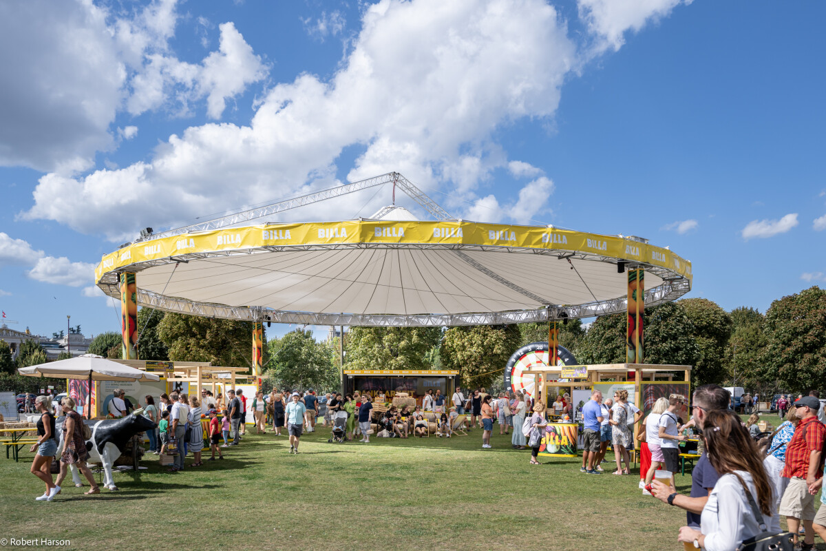 Menschen auf einem Rasenplatz bei einem Outdoor-Event an einem sonnigen Tag. Eine große, runde Struktur mit gelber Markise bietet Schatten. Stände mit verschiedenen Angeboten sind sichtbar, und im Hintergrund stehen Bäume.