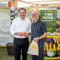 Zwei Männer stehen vor einem Billa-Stand auf einer Veranstaltung, lächeln in die Kamera und halten Obst und eine Billa-Tasche. Im Hintergrund sind Banner mit Informationen zur Tierwohl-Initiative zu sehen.