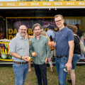 Drei Männer stehen auf einer Wiese und halten Äpfel in der Hand. Hinter ihnen ist ein BILLA-Stand mit Menschen im Hintergrund.