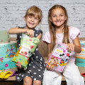 Zwei lachende Mädchen sitzen vor einer weißen Backsteinwand. Eines hält eine Packung Apfelchips und das andere eine Packung Cornflakes, beide mit Disney-Motiven. Um sie herum sind weitere essbare Produkte und Küchenrollen mit ähnlichen Designs.