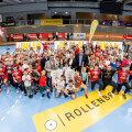 Eine große Gruppe von Menschen, darunter viele Kinder in Sportkleidung, posiert in einer Sporthalle. Sie halten Schilder und sind von Sponsorenlogos umgeben. Einige Erwachsene stehen in der vorderen Reihe. Alle scheinen fröhlich zu sein.