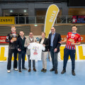 Sechs Personen stehen in einer Sporthalle und halten ein T-Shirt. Sie lächeln, die Halle ist mit Bannern wie "ROLLENBAU" und "BILLA" dekoriert.