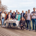 Eine Gruppe von Menschen steht auf einer Holzterrasse vor einem Tisch mit Kuchen und Gebäck. Sie posieren für ein Gruppenfoto. Im Hintergrund sind Bäume und ein bewölkter Himmel zu sehen.