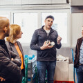 Eine Gruppe von fünf Personen steht in einer industriellen Umgebung. Ein Mann in der Mitte trägt eine Mütze und hält Gläser in der Hand, während er mit den anderen spricht. Zwei Männer zur Rechten hören aufmerksam zu, und eine Frau zur Linken schaut ebenfalls zu. Ein Mann im Vordergrund ist von hinten zu sehen. Im Hintergrund sind Fenster und Regale.