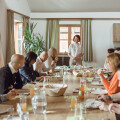 Eine Gruppe von Personen sitzt um einen langen Holztisch in einem hellen Raum. Sie essen und hören einer stehenden Person zu, die spricht. Auf dem Tisch stehen Gläser, Teller und Saftflaschen. Im Hintergrund sind große Fenster und Pflanzen zu sehen.
